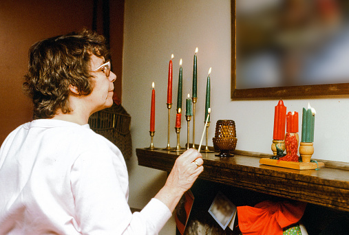 An older woman lights a group of red and green candles on the fireplace mantle.  It is time for the Christmas holidays. The horizontal image is simple, yet festive. A flash bounce is used to light the evening image.