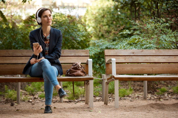 Woman listening to music on bench in park Full length of woman listening music with headphones and mobile phone. Thoughtful female is sitting on wooden bench in park. She is relaxing while looking away outdoors. bench stock pictures, royalty-free photos & images