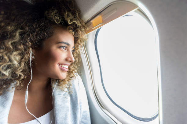 Woman looking through window on airplane Confident young mixed race woman smiles while looking through window on aircraft. She is wearing earbuds. plane window seat stock pictures, royalty-free photos & images