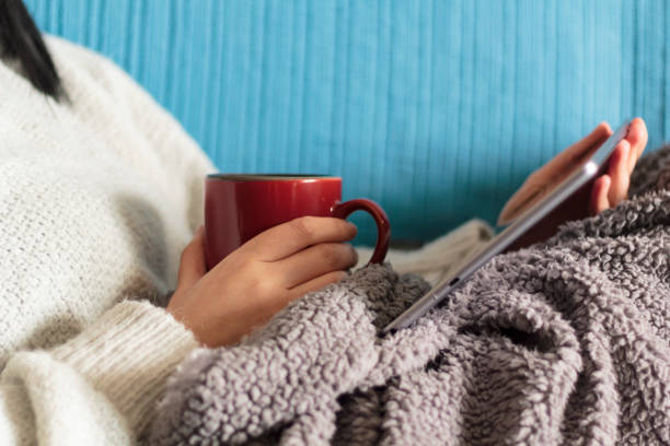 Woman lying on sofa with blanket watching a movie on tablet and holding a mug stock photo