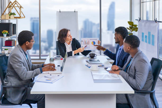 Woman manager is showing annual report chart to her African American colleagues in the executive meeting for next year plan with city skyline background for global business and investment concept stock photo