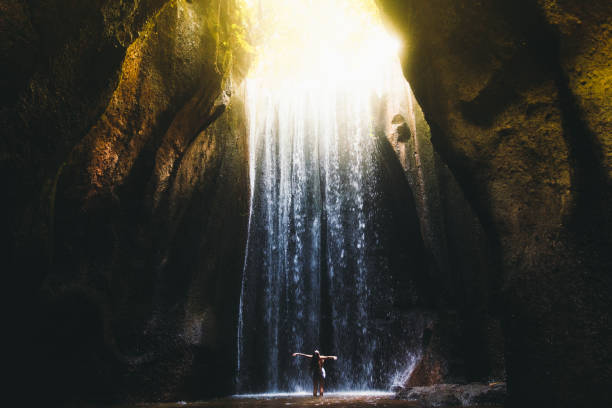 Woman meets sunrise in the cave under the big waterfall on Bali island, Indonesia Young woman feeling awe and happiness getting wet staying under the huge waterfall hidden in the cave during bright summer sunny sunset on Bali, Indonesia cave photos stock pictures, royalty-free photos & images