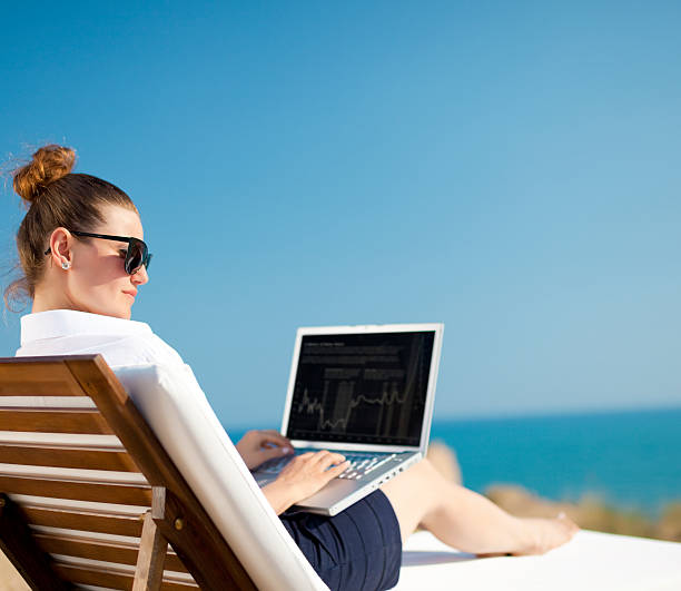 Woman on wooden lounge chair working on laptop at the beach  stock photo