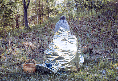 Woman person is lost and sit in wild forest in cold day, using first aid emergency blanket to prevent hypothermia and body heat loss. Emergency blanket concept.