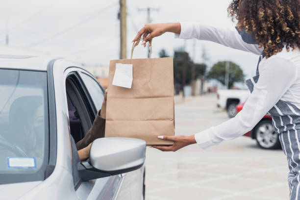 Woman picks up take out order A waitress hands a bag of food to a female customer who has ordered food from the restaurant. The customer is picking up the food from curbside pickup. curbsidepickup stock pictures, royalty-free photos & images