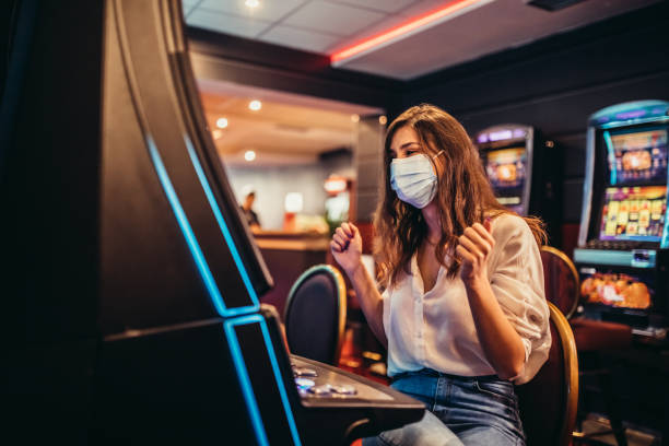 Woman playing slot machine in casino Beautiful young woman with protective face mask playing slot machine in casino. casino stock pictures, royalty-free photos & images