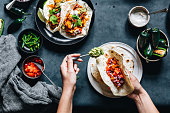 Point of view shot of a woman preparing tasty vegan tacos in kitchen. Female hands put fillings in tacos.