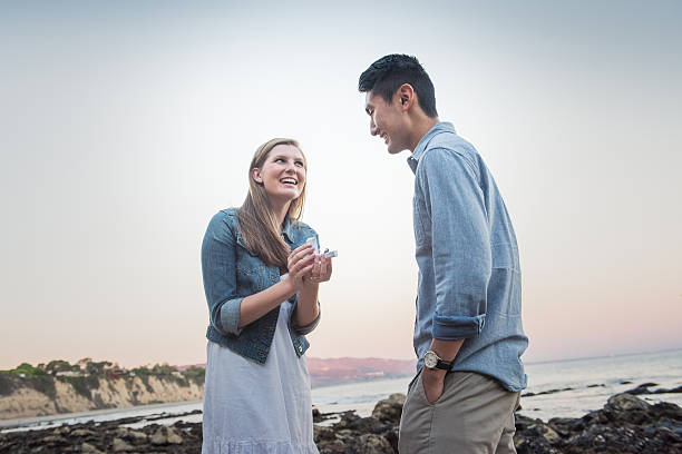 Woman Propsing to Her Boyfriend at the Beach A true role reversal. Caucasian young woman proposing with a diamond ring to his Japanese boyfriend at the beach during sunset. custom ring couple stock pictures, royalty-free photos & images