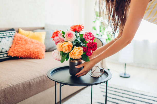 Woman puts vase with flowers roses on table. Housewife taking care of coziness in apartment. Interior and decor stock photo