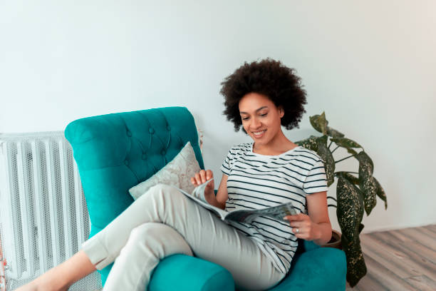 Woman reading a magazine Beautiful young mixed race woman sitting in an armchair, reading a magazine and relaxing at home curley cup stock pictures, royalty-free photos & images