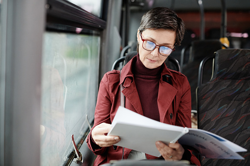 Portrait of elegant mature woman reading book on bus while traveling by public transport in city, copy space