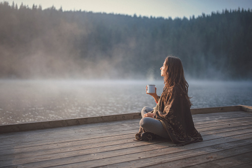 Side view of woman sitting on wooden pier in nature, she looking the sunrise and drinking coffee.