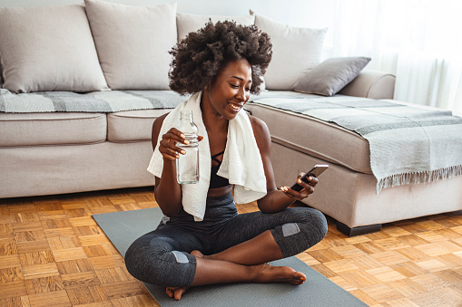 Woman resting after home workout with water and phone. Young sporty woman after practicing yoga, break in doing exercise, relaxing on yoga mat, texting on the cell phone, holding smartphone, friendly smiling reading message