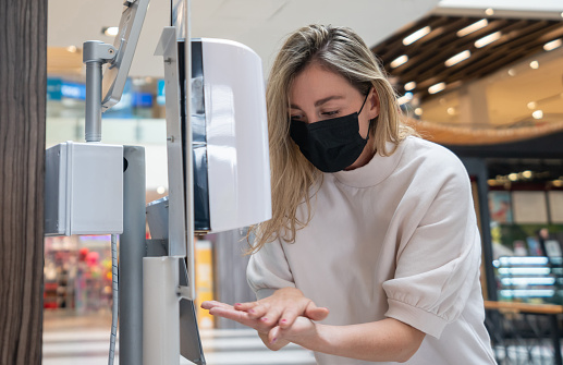Woman shopping at the mall during the pandemic wearing a facemask and disinfecting her hands at a hand sanitizer station - COVID-19 lifestyle concepts