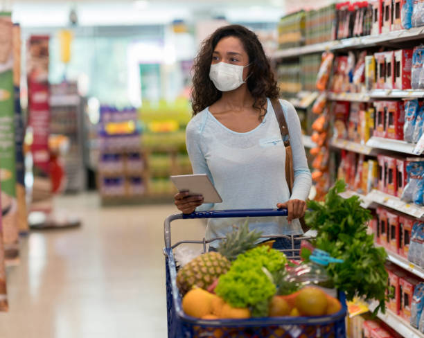 Woman shopping at the supermarket wearing a facemask Woman shopping at the supermarket wearing a facemask to avoid the coronavirus while following a list on her tablet computer â COVID-19 lifestyle concepts shopping list stock pictures, royalty-free photos & images