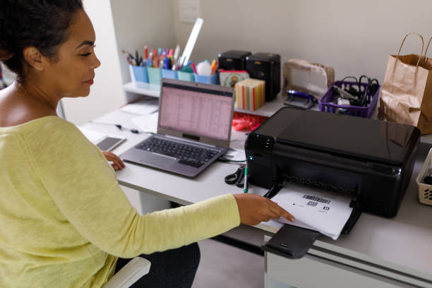 donna seduta alla scrivania a raccogliere la carta del codice identificativo dalla stampante - lavorare da casa foto e immagini stock