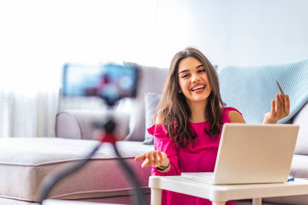 Woman sitting beside desk with laptop while filming her broadcast Happy girl at home speaking in front of camera for vlog. Young woman working as blogger, recording video tutorial for Internet. Young female blogger with laptop recording video at home"r"n influencer stock pictures, royalty-free photos & images