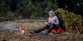 Woman reading a book while sitting next to a campfire and a makeshift shelter on a woodland plain.