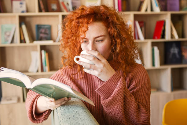 mujer sentada en el sofá con taza de café en la librería leyendo un libro - curley cup fotografías e imágenes de stock