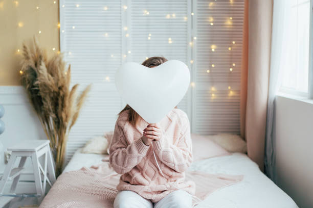 A woman sitting on the bed  hid her face behind an inflatable balloon in the shape of a heart. stock photo