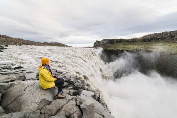 Woman sitting on the cliff edge next to Dettifoss waterfall in Vatnayokull national park, Iceland Woman in yellow raincoat sitting standing on the cliff edge next to Dettifoss waterfall in Vatnayokull national park, Iceland. iceland dettifoss stock pictures, royalty-free photos & images