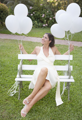 Woman Sitting Outdoors On Bench With Balloons Tied To It Smiling