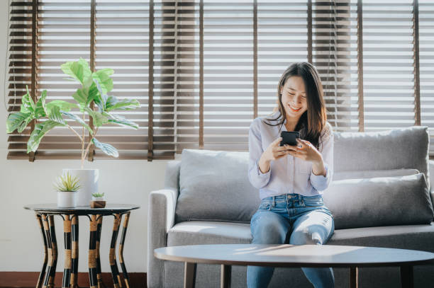woman smiling and texting on the smartphone sitting on sofa Happy Asian woman smiling and texting on the smartphone sitting on sofa i lving room at her home asian woman using phone stock pictures, royalty-free photos & images