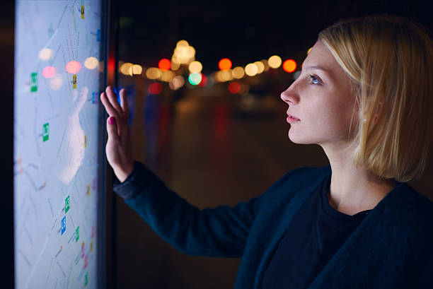 Woman standing front big digital screen with basic city map Gorgeous young female touching sensitive display of modern smart city bus stop for check her location into the night touch screen stock pictures, royalty-free photos & images
