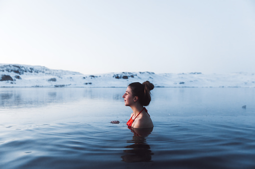 Young woman in red swimsuit bathing in picturesque hot swimming pool enjoying the view of snowy mountain peaks in North Iceland