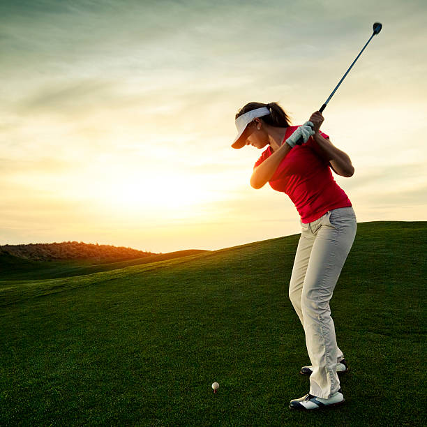 Woman swings golf club on a grassy hill at sunset stock photo