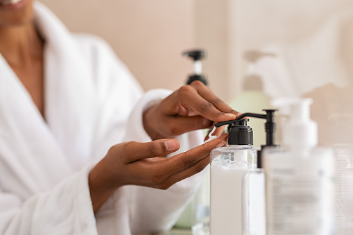 African woman hands using cosmetic liquid soap in bathroom. Close up of girl black hands in bath robe using body lotion dispenser after shower. Black girl putting pomade on hand from pump.