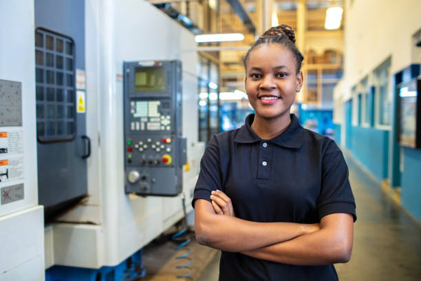 Woman technician standing in factory shop floor Portrait of a young woman technician standing in factory shop floor. Female in uniform looking at camera with her hands folded. cnc machine stock pictures, royalty-free photos & images
