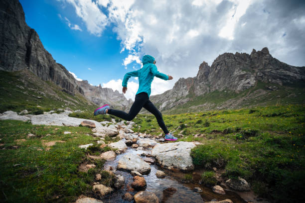 Woman trail runner jumping over samll river on beautiful mountains stock photo