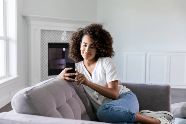 Woman uses smartphone in new home Relaxed young woman smiles while video chatting with friend on smartphone. She is relaxing on the sofa in her living room. mobile real estate stock pictures, royalty-free photos & images