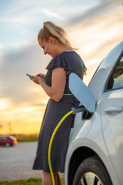 kvinna som använder telefon och väntar på elbil att ladda vid solnedgången - electric car woman bildbanksfoton och bilder