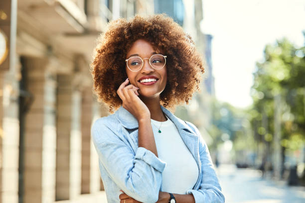 Woman using smart phone at sidewalk in city Smiling young woman talking on smart phone. Beautiful female is wearing eyeglasses on sidewalk. She is having frizzy hair. eyewear stock pictures, royalty-free photos & images