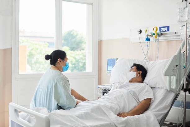 Woman Visiting Male Patient in Hospital Ward Woman visiting male patient in hospital ward. Female is sitting by man lying on bed. They are at hospital during COVID-19 epidemic. visit stock pictures, royalty-free photos & images