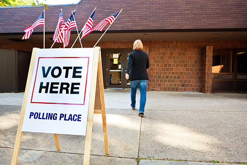 Unidentifiable woman voter entering a voting polling place for USA government election. Rear view shows her walking briskly in blurred motion by a sign decorated with American flags. For concepts of one person one vote, voter registration, voting booths, presidential and legistlative national and local decisions, democracy, and civic responsibility.