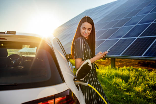 Woman waiting for electric car to charge and solar panels in background Waist up shot of a smiling young woman with black hair waiting for an electric car to charge and using a mobile phone with solar panels visible in the background electric car photos stock pictures, royalty-free photos & images