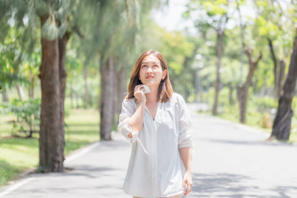 Woman walking in the park having sunstroke in summer hot weather, Young pretty girl drying sweat using a wipe on a warm summer day in a park stock photo