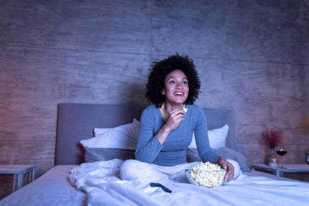 Woman watching comedy on TV Beautiful mixed race woman wearing pajamas sitting on bed, eating popcorn and watching a comedy movie on TV, relaxing at home late at night watching stock pictures, royalty-free photos & images