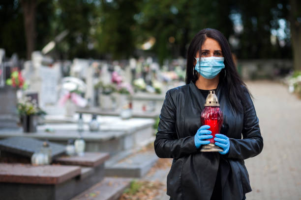 a woman wearing a protective mask against the coronavirus covid-19 sars-cov-2 holds a candle in her hand and visits her relatives at the cemetery. lockdown cemetery - covid cemiterio imagens e fotografias de stock