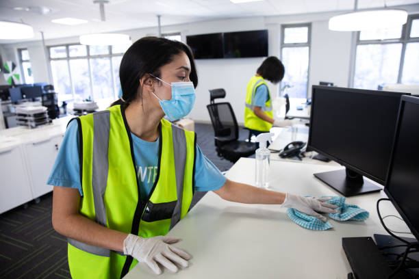 Woman wearing hi vis vest and face mask cleaning the office using disinfectant Mixed race woman and colleague wearing hi vis vests, gloves and face masks sanitizing an office using disinfectant. Hygiene in workplace during Coronavirus Covid 19 pandemic. office cleaning stock pictures, royalty-free photos & images