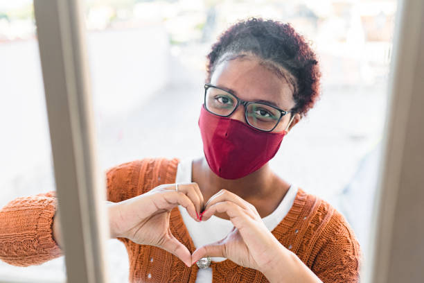 Woman wearing mask and making heart sign through window Smile behind the mask during the Covid-19 pandemic. resilience stock pictures, royalty-free photos & images