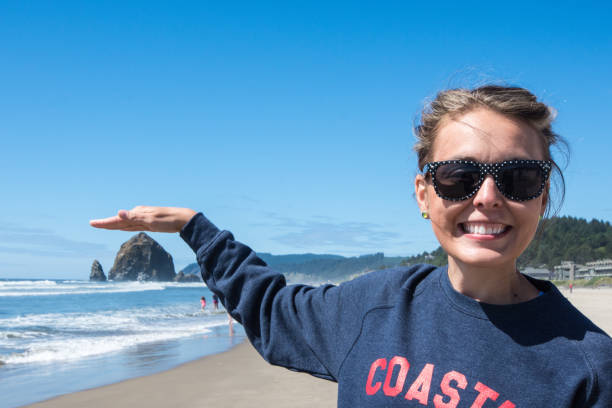 la donna che indossa occhiali da sole finge di toccare una roccia haystack a cannon beach, oregon, sulla spiaggia in una giornata di sole. visualizzazione prospettiva forzata - forced perspective foto e immagini stock