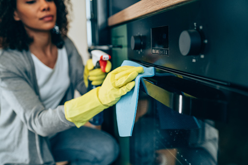 Shot of young woman cleaning the outside of an oven. Woman cleaning with spray disinfectant and gloves at home.