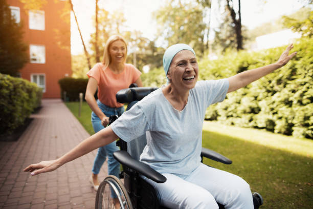 A woman with cancer is sitting in a wheelchair. She walks on the street with her daughter and they fool around. A woman with cancer is sitting in a wheelchair. She walks on the street with her daughter and they fool around. They are fun and they laugh. They walk in the courtyard of the clinic. cancer illness stock pictures, royalty-free photos & images