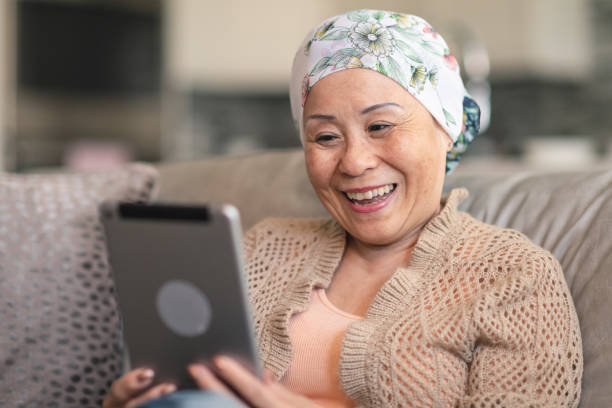 Woman with cancer video chats with friends on digital tablet A senior adult woman of Chinese descent has cancer. The happy woman is using a digital tablet to video message friends and family. She is spending time at home. The woman is smiling at the screen. She is wearing a bandana to hide her hair loss from chemotherapy treatment. cancer illness stock pictures, royalty-free photos & images