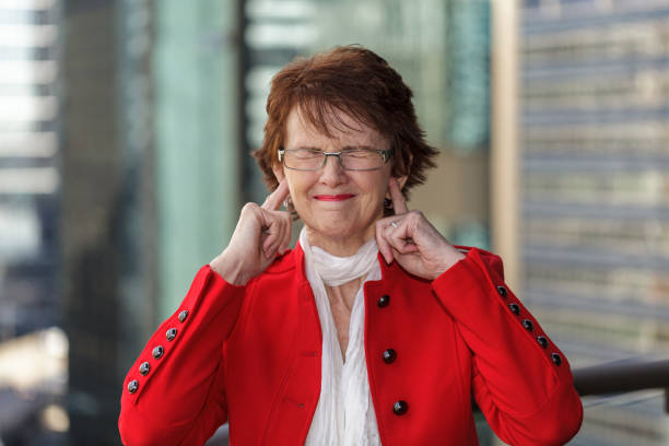 Woman with eyes closed and fingers in her ears. Businesswoman in red jacket, with out of focus office buildings in the background. Fingers in Ears stock pictures, royalty-free photos & images