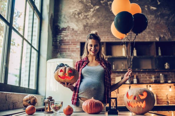Woman with Halloween pumpkin. Happy Halloween! Attractive young pregnant woman is preparing to Halloween on kitchen. Beautiful woman with pumpkin and air balloons in hands. big smile emoji stock pictures, royalty-free photos & images